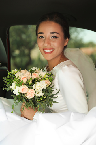 Young bride wearing wedding dress with beautiful bouquet in automobile