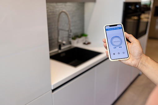An over the shoulder view of an unrecognisable mans hand holding a fridge thermostat app on his phone and being energy efficient in his smart home.