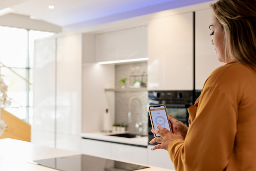 An over ths shoulder view of a young woman controlling the temperature of her home using her smart phone. She is is her modern kitchen in Hexham in the North East of England.