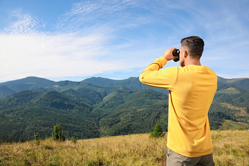 Man with binoculars in mountains on sunny day, space for text