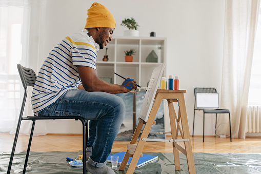 Black man sitting on the chair and painting on the white canvas in the living room