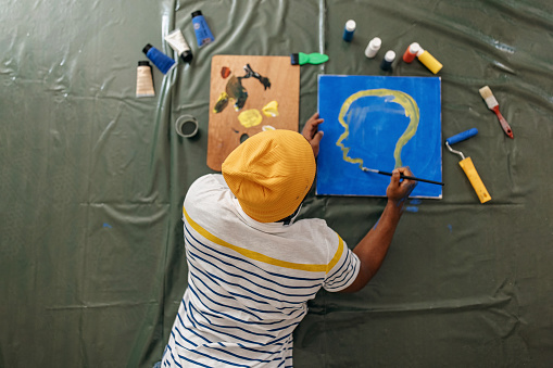 Black man wearing yellow hat painting on the canvas, shot from above