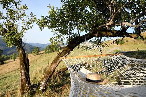 Comfortable net hammock with hat and book in mountains on sunny day