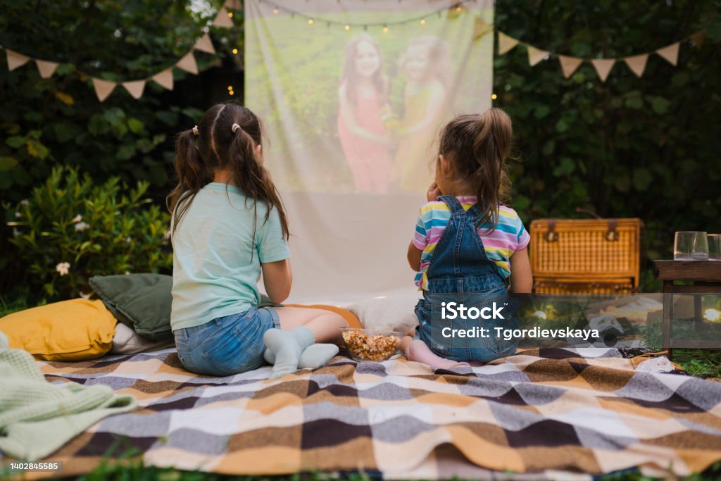 Backyard Family outdoor movie night with kids. Sisters spending time together and watching cimema at backyard. DIY Screen with film. Summer outdoor weekend activities with children. Open air cinema. Movie Theater Stock Photo