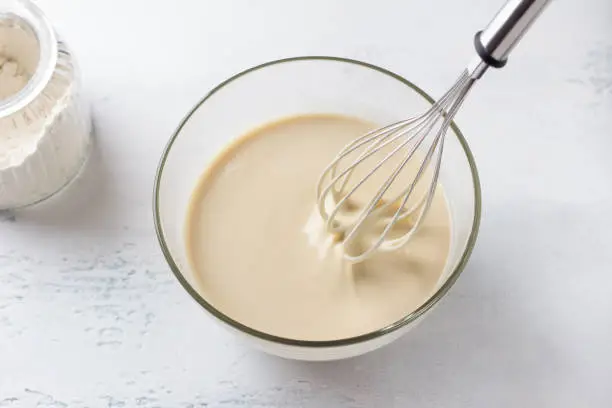 A glass bowl of dough for Finnish pancakes with a whisk and a jar of flour on a light gray background, top view. Cooking delicious homemade food