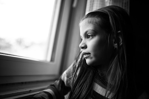 Black and white image of cute little girl who is pensive and looking through window.
