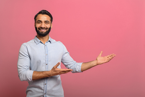 Handsome Indian guy points palms at empty copy space isolated on pink background. Cheerful multiracial bearded man in casual jeans shirt presenting novelty, advertising concept