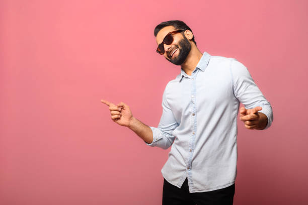 Overjoyed Indian man wearing sunglasses dancing, makes movements to music Overjoyed Indian man wearing sunglasses dancing, makes movements to music, smiles positively, being in high spirit. Carefree hispanic guy in blue jeans shirt dances isolated on pink fashion model men male sunglasses stock pictures, royalty-free photos & images