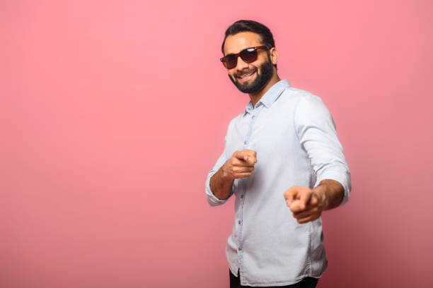 Handsome smiling young indian man in jeans shirt pointing fingers at you Handsome smiling young indian man in jeans shirt pointing fingers at you standing isolated on pink background, cheerful multiracial guy indicating at the camera arabian sunglasses stock pictures, royalty-free photos & images