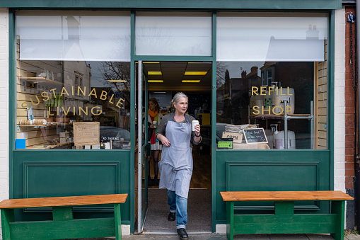 Woman working at a cafe/store that promotes sustainable living in the North East of England. The store has refill stations to reduce plastic and food waste. She is wearing an apron and walking out the door to enjoy her break with a coffee.