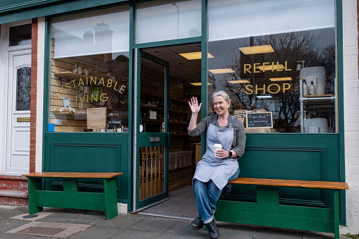 Woman working at a cafe/store that promotes sustainable living in the North East of England. The store has refill stations to reduce plastic and food waste. She is wearing an apron and sitting down outside the front of the store to enjoy her break with a coffee.