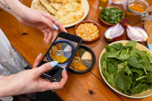 A close-up shot of an unrecognisable young woman maintaining her sustainable living by preparing a vegan meal with a collection of fresh spices and ingredients in the kitchen in the North East of England. She is taking a picture of the spices with her smartphone.