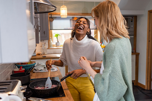 Two friends maintaining their sustainable living by preparing a vegan meal in the kitchen in the North East of England. One young woman is cooking chopped onions in the pan while her friend stands with her holding a bottle of olive oil.