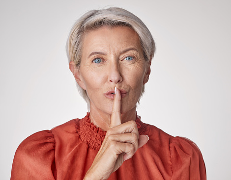 One mature caucasian woman gesturing with a finger on her lips to be quiet and keep a secret against a grey copyspace background. Ageing woman hushing for silence and whispering a scandal