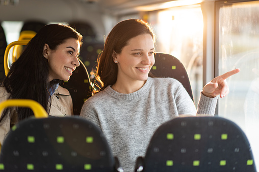 Two happy female friends looking at something and pointing whit finger while traveling by bus