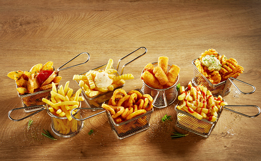 From above of assorted types of French fries topped with sauces served in metal meshes on wooden table in light kitchen