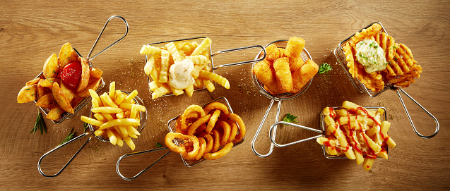 Top view of various delicious French fries with sauces served in metal meshes on wooden table in light room