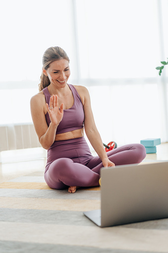 Woman practicing yoga with trainer via video conference. Beautiful young woman doing workout at home. Woman doing exercises while watching workout video. Young woman sitting in front of her laptop and watching online sport lessons. Woman exercising and watching online fitness live streaming classes