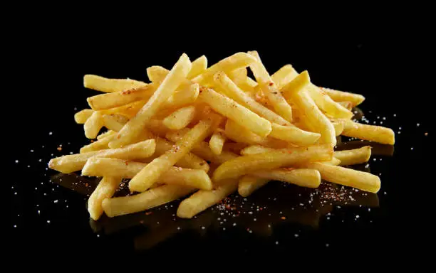 From above of delicious savory crunchy French fries with salt and red pepper on black glossy table