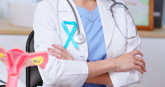 close up female doctor wear white coat with teal ribbon arm crossed in front of her chest - uterus model on table