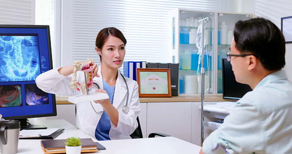 attractive young brunette ponytail female asian doctor wear white coat and stethoscope explaining colon model to sick male patient at clinic- an xray film display on computer