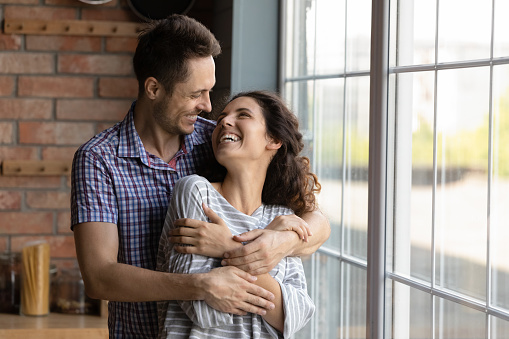 Affectionate happy young man cuddling smiling attractive wife, standing near huge window in own apartment. Smiling loving millennial latin family couple showing tender feelings, communicating indoors.