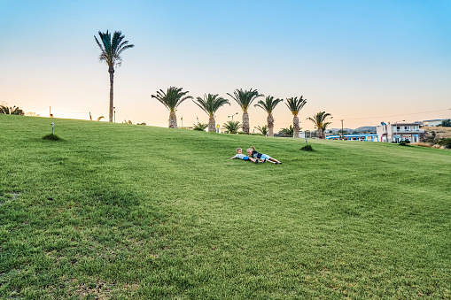 Brothers lie on soft green grass on lawn against palm trees near hotel building. Preschooler and schooler relax at twilight on summer vacation