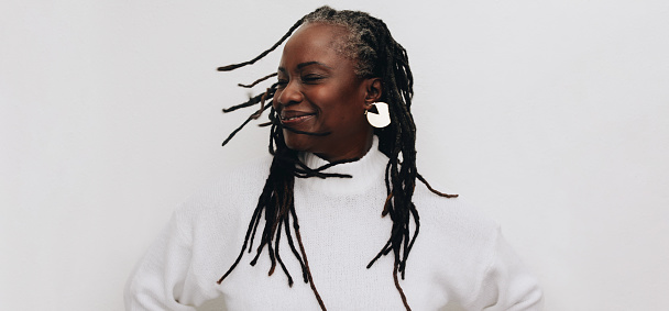 Happy mature woman whipping her dreadlocks confidently while standing against a white background. Body positive black woman embracing her natural hair.
