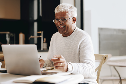 Happy senior businessman writing notes during a virtual meeting in his home office. Mature businessman having a video conference with his colleagues while working from home.