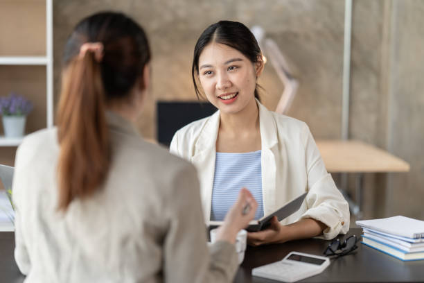 Young beautiful asian business woman is talking to her colleague, while sitting at the desk in a modern office. Concept of business success. Young beautiful asian business woman is talking to her colleague, while sitting at the desk in a modern office. Concept of business success. HR stock pictures, royalty-free photos & images