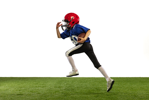 Dynamic portrait of little boy, beginner player of american football training isolated on white background with green grass flooring. Concept of sport, movement, achievements. Copy space for ad