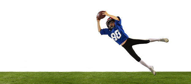 Workout. Sportive little boy in sports uniform and equipment playing american football isolated on white background with green grass flooring. Concept of sport, ad, movement, achievements.