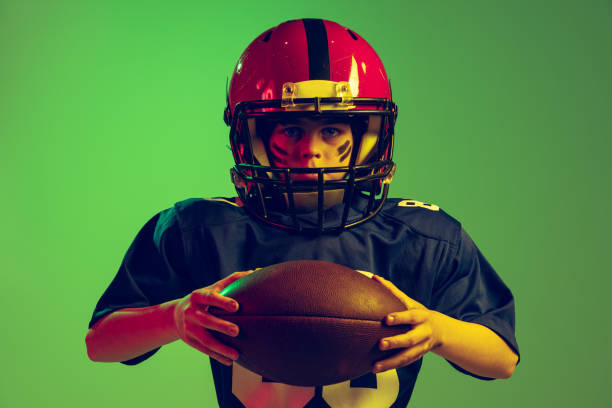 学齢期の男の子、スポーツユニフォームを着た初心者アメリカンフットボール選手、ネオンライトの緑色の背景に隔離されたヘルメットのクローズアップポートレート。 - football helmet american football green american football uniform ストックフォトと画像
