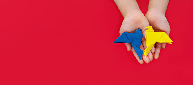 two doves of peace made of origami paper in yellow and blue of colors of Ukrainian flag in children's hands on red background