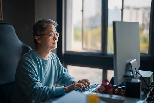 asian chinese mature man work from home typing and looking at computer at his home office