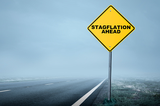 A road sign at the side of a rural road warns of stagflation ahead as the road descends into a shroud of fog.