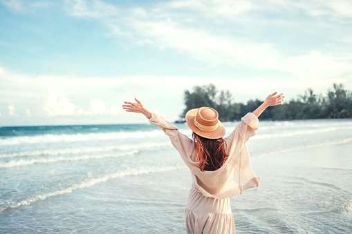 Summer beach vacation concept, Young woman with hat relaxing with her arms raised to her head enjoying looking view of beach ocean on hot summer day, copy space.