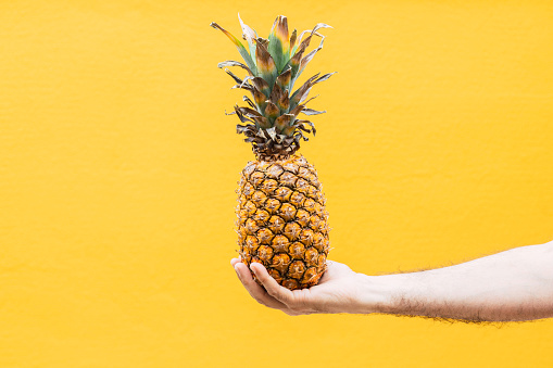Unrecognizable person holding a pineapple in front of a yellow wall.\nConceptual of summer, healthy eating