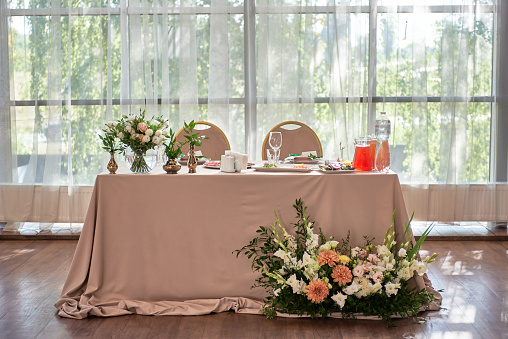 Wedding presidium in restaurant, copy space. Banquet table for newlyweds with flowers, greenery, candles and pink cloth. Lush floral arrangement. Luxury wedding decorations