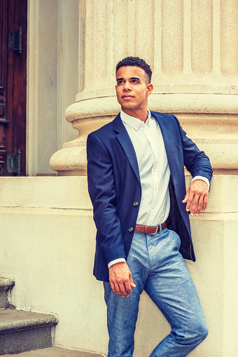 African American Businessman working in New York. Wearing blue blazer, white shirt, gray pants, a black college student standing by column outside on campus, looking up.