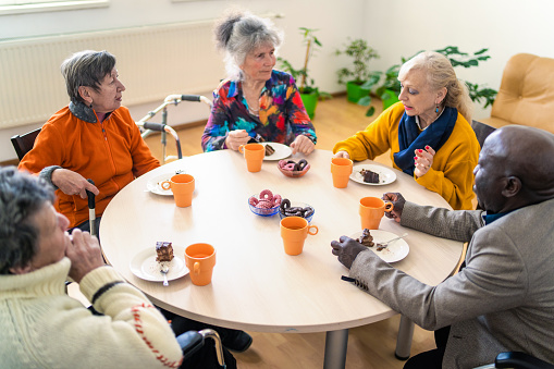 A high angle view of five residents in a care home, sitting around a table drinking tea and eating cake for a morning snack