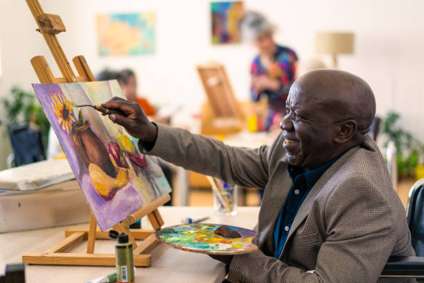 Retired pensioner painting for therapy A senior African-American man is painting a picture using a paintbrush and palette for his composition on the canvas, behind him other members of the community are also painting accessibility for persons with disabilities photos stock pictures, royalty-free photos & images