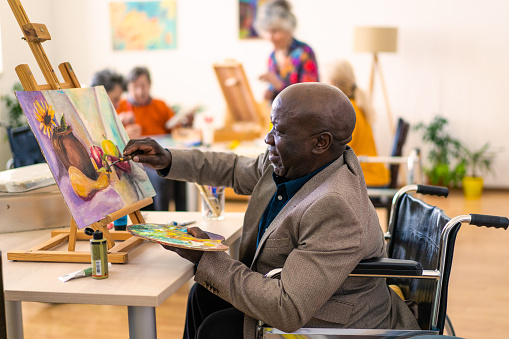 Sitting in a wheelchair at the local elder care centre, a mature man is painting a picture during an art therapy group