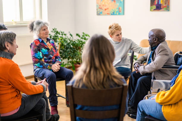 A caring clinician offers sympathy to an elderly man A female psychoanalyst placing a comforting hand onto a mature man's shoulder while conducting a therapy meeting in a bright and airy room with senior residents of a aged care home group therapy stock pictures, royalty-free photos & images