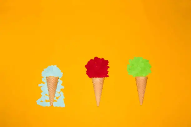 Photo of three ice cream cones, one with a green paper ball, another with red, and a third with melted blue. creative summer design on a yellow background