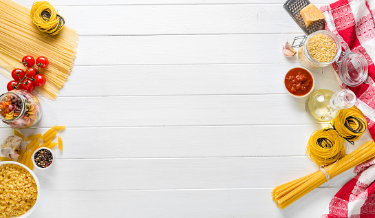 Top view of Italian raw spaghetti and pasta nests with olive oil, cheese, garlic and tomatoes on white wooden background with free space for text or recipe