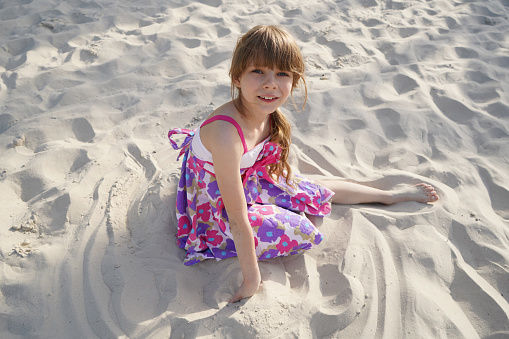 A 7-8-year-old girl with dark hair is sitting in white sand in a sundress with a big flower