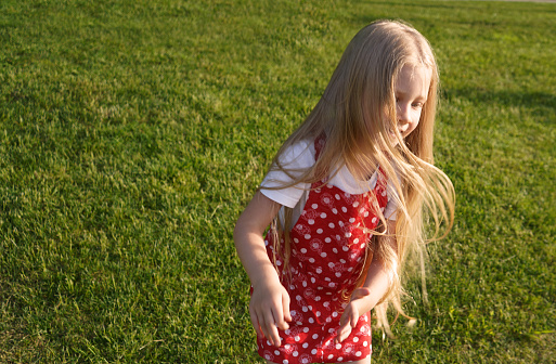 A happy 5-year-old European girl with blonde hair in a red sundress and a white T-shirt jumps on the spot on the grass