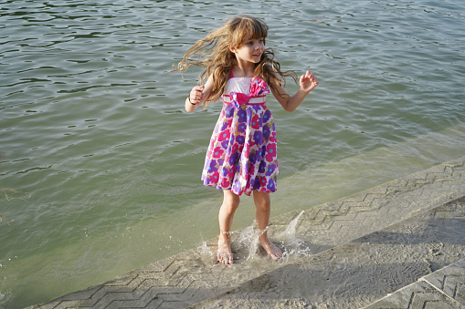 A happy 7-year-old girl with dark hair in a colored sundress jumps barefoot on the shore of the lake at sunset, splashes of water fly
