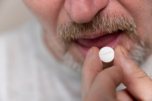 Mature man taking medication. Lower part of the male face. Man places a round white pill on his tongue with his hand. Male with stubble. Gray hair on his beard and mustache. Close-up. Selective focus